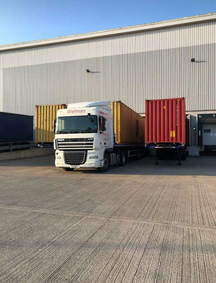 Flat bed lorry loaded with yellow shipping container parked in front of warehouse